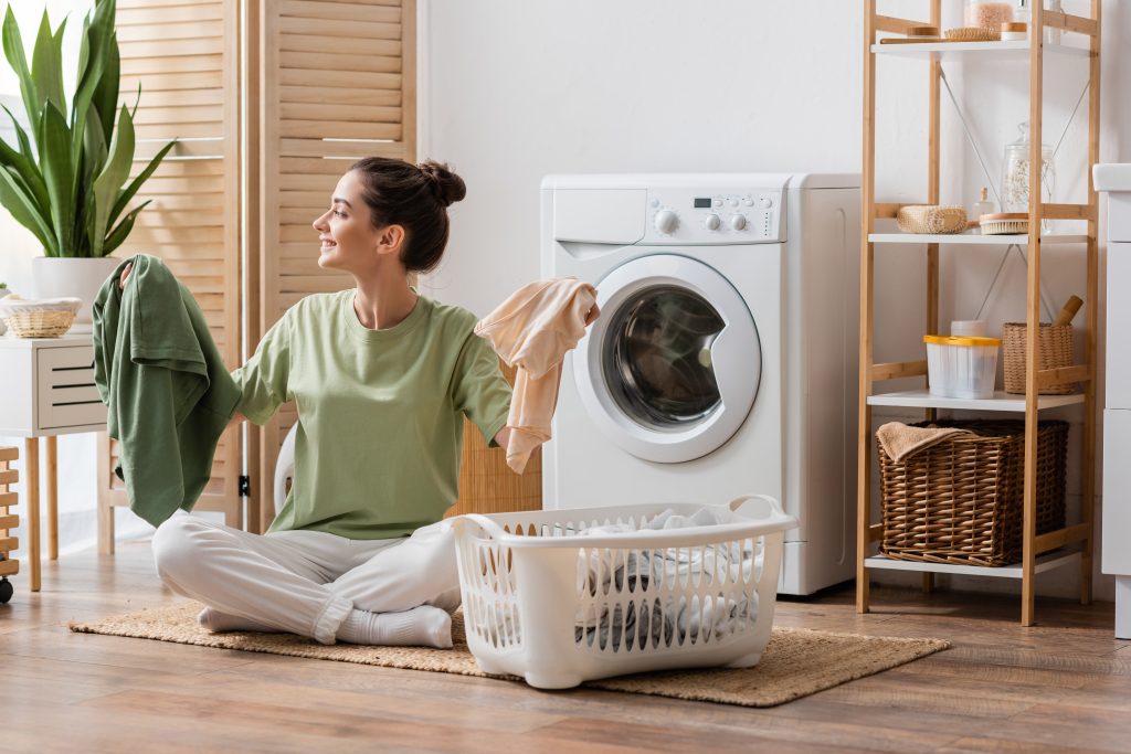 Side,View,Of,Cheerful,Brunette,Woman,Holding,Clothes,Near,Basket